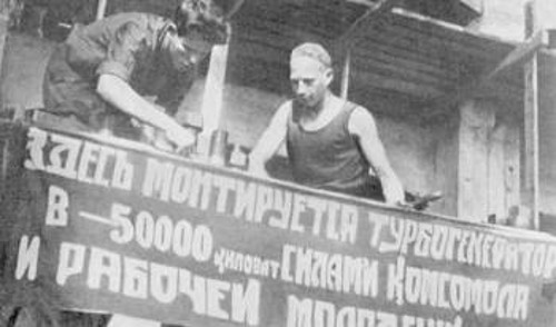 «Εδώ συναρμολογείται τουρμπογεννήτρια 5.000 κιλοβάτ, από τις δυνάμεις της ΚΟΜΣΟΜΟΛ και της εργατικής νεολαίας», γράφει το αναρτημένο πανό σε σοβιετικό εργοστάσιο