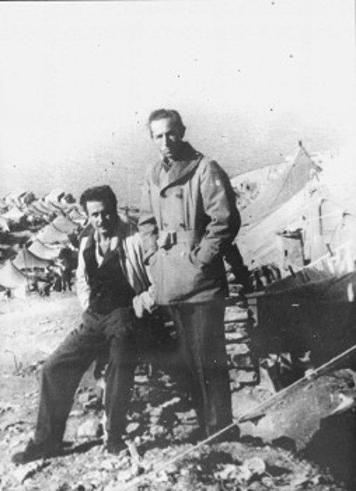Ο Γιάννης Ρίτσος με τον σύντροφό του, ηθοποιό Μάνο Κατράκη, μπροστά στη σκηνή τους, το 1949, στη Μακρόνησο. Πίσω τους ο φωτογραφικός φακός έχει συλλάβει τα καταλύματα κι άλλων εξόριστων