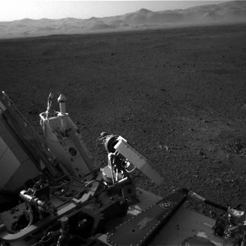 Στη φωτογραφία φαίνεται ένα μέρος του Curiosity και των συστημάτων του, η σχεδόν επίπεδη περιοχή γύρω από το σημείο προσεδάφισης και στο βάθος βουνά του Αρη