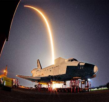 Σε πρώτο πλάνο ένα παροπλισμένο Διαστημικό Λεωφορείο της NASA και στο βάθος το αποτύπωμα στο φωτογραφικό φακό (με μακράς διάρκειας λήψη) από την εκτόξευση του ιδιωτικού διαστημικού σκάφους