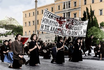 Αθήνα 4 Δεκέμβρη 1944. Χιλιάδες λαού στην κηδεία των θυμάτων της προηγούμενης μέρας. Ανάμεσά τους τα μαυροφορεμένα κορίτσια κρατώντας το πανό που έγραφε: «Οταν ο λαός βρίσκεται μπροστά στον κίνδυνο της τυραννίας διαλέγει ή τις αλυσίδες ή τα όπλα - ΕΑΜ»